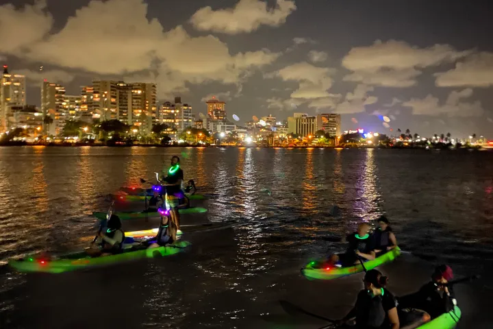 People kayaking at night with neon lights, city skyline in background.