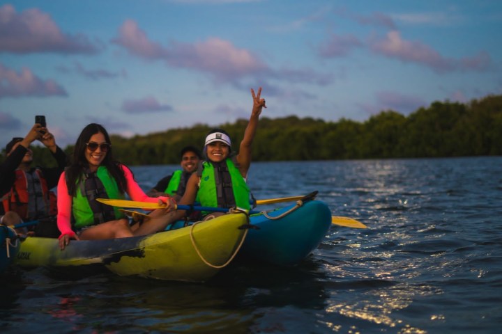 Group kayaking on a lake at sunset, wearing life jackets, one person flashing a peace sign.