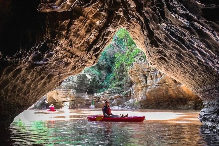 Kayakers in a cave with sunlight illuminating rock walls and green water.