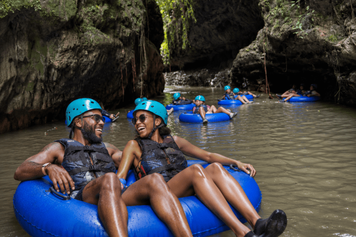 People in blue helmets tubing on a river through a rocky, lush canyon.