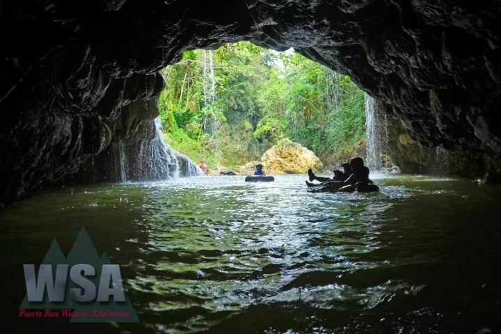 People tubing through a cave with a waterfall, surrounded by lush greenery.