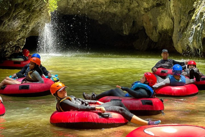 People in inflatable tubes near a rocky cave entrance with a small waterfall.