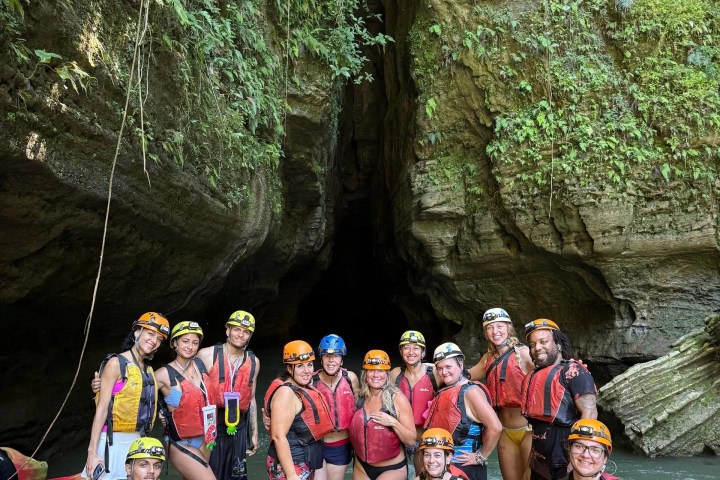 Group in helmets and life vests pose at rocky river entrance of lush canyon.