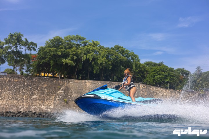 Person riding a blue jet ski on water near a stone wall and trees.